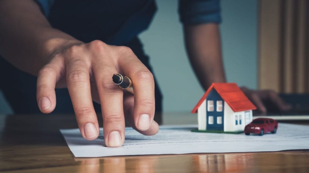 Mão com caneta sobre documento de compra de imóvel ao lado de maquete de casa